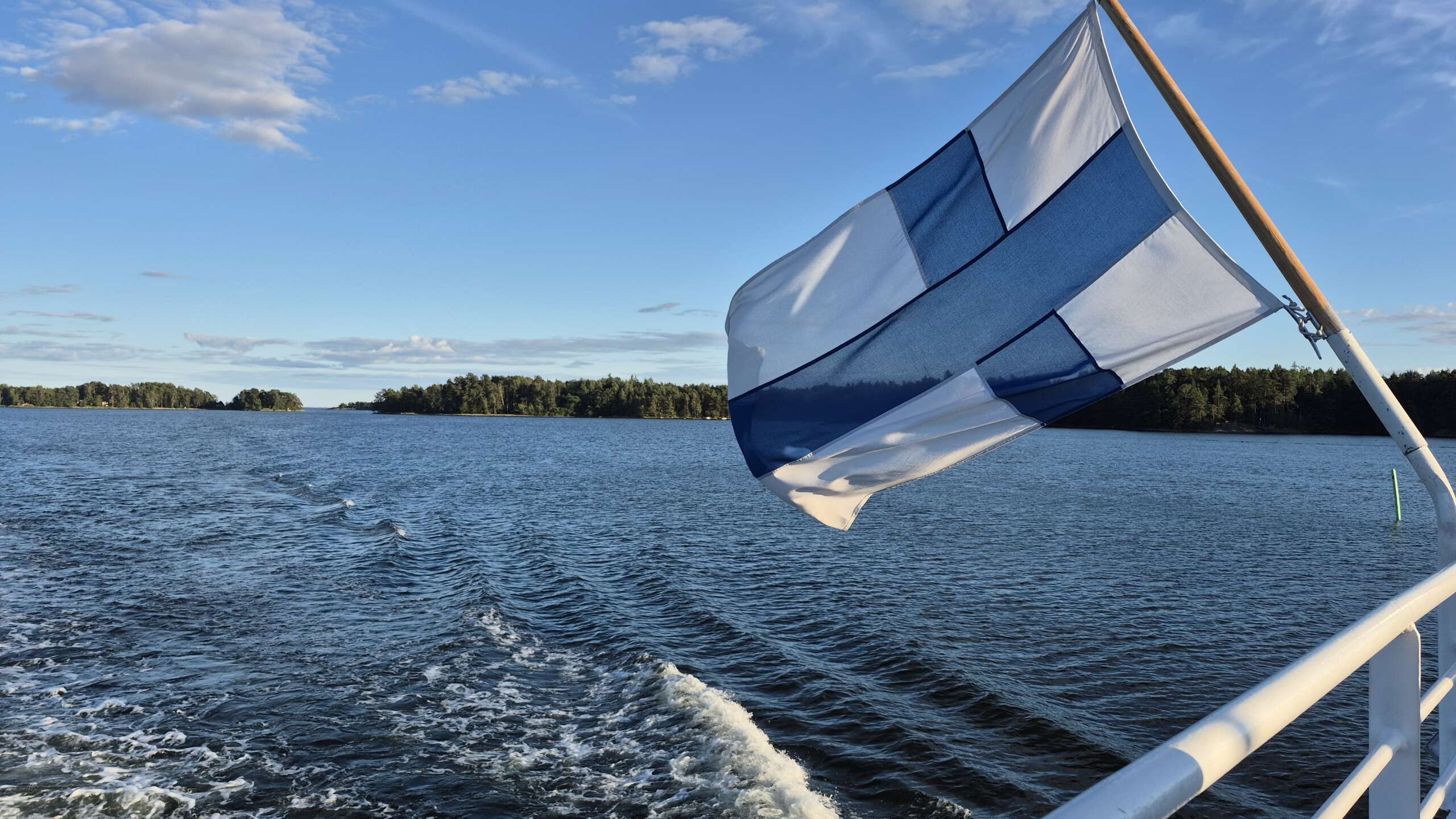 Eine finnische Flagge weht am Heck eines Bootes auf ruhigem Wasser, mit bewaldeten Inseln und einem teilweise bewölkten Himmel im Hintergrund.
