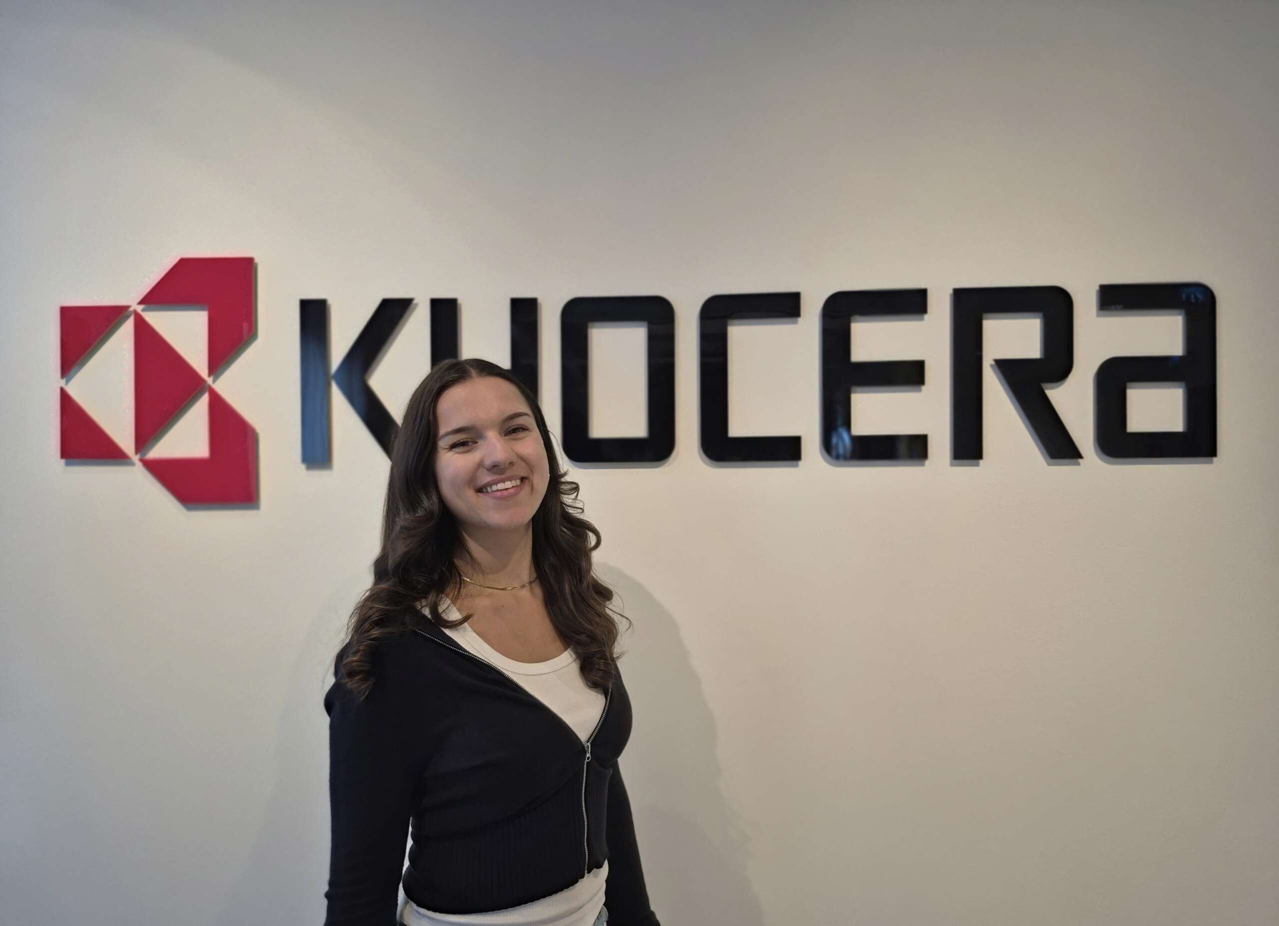 Eine Frau lächelt vor einer Wand, auf der das Kyocera-Logo und der Name in fetten schwarzen Buchstaben stehen.