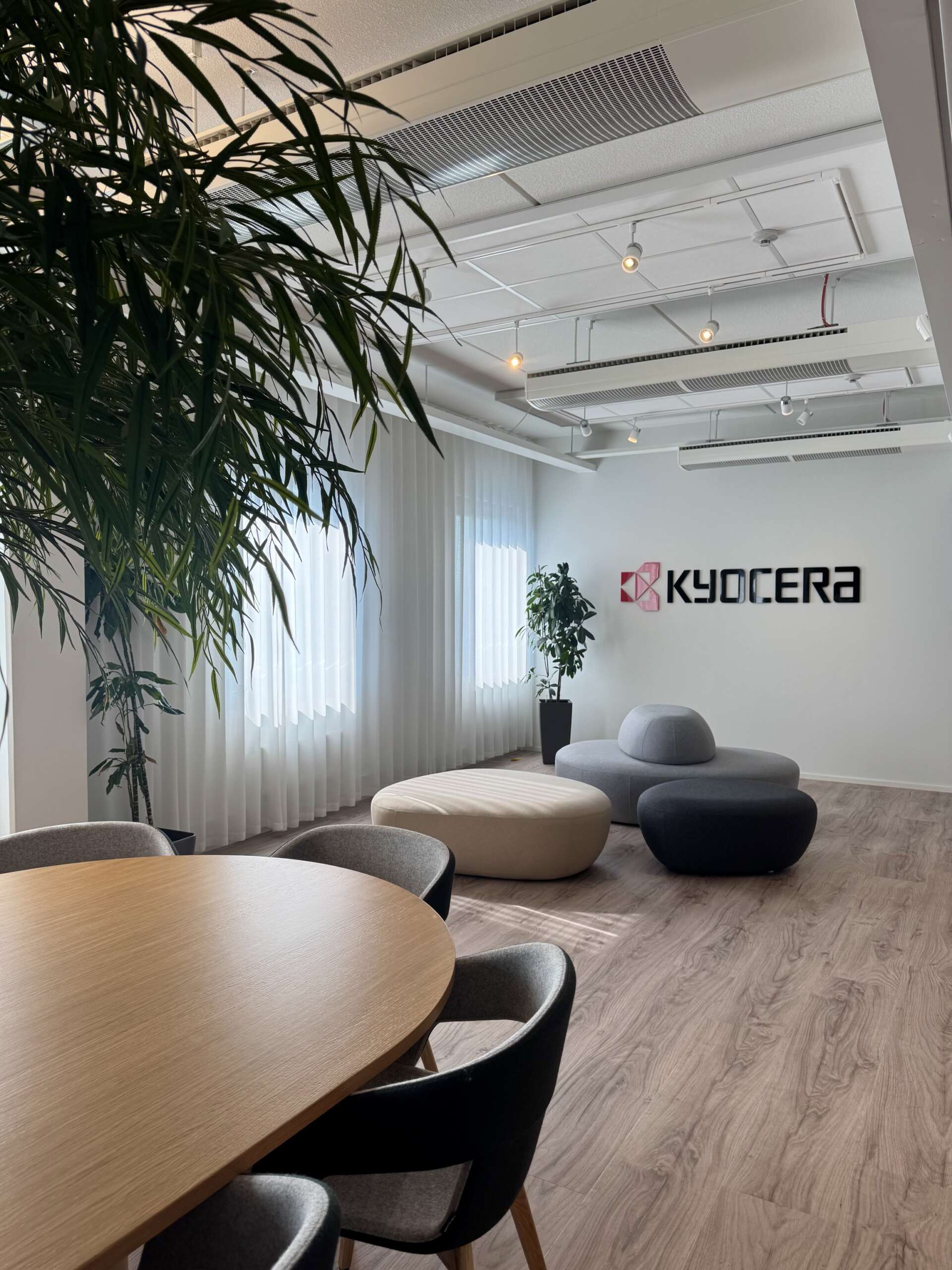 Büro von KYOCERA mit Rundsitzgruppe, Holztisch, Stühlen, großen Fenstern, Topfpflanzen und einem KYOCERA-Logo an einer weißen Wand.